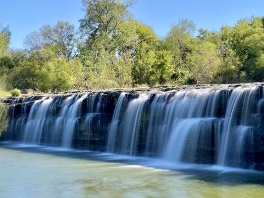 A scenic view of the wide, natural limestone waterfall at Marion Sansom Park near Azle, Texas, with water cascading over rocky ledges into a pool surrounded by lush green forest.