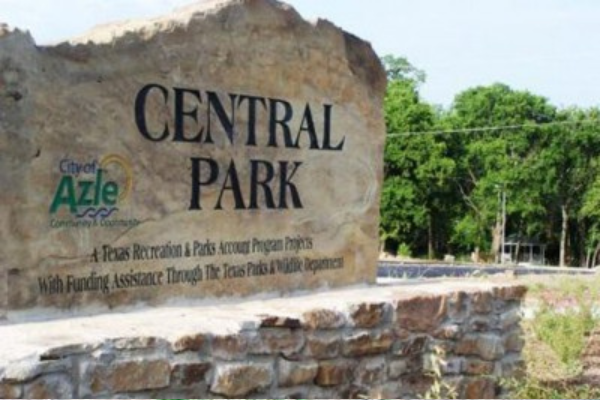 A large stone monument sign at the entrance of Central Park in Azle, Texas, featuring the city logo and park name against a backdrop of lush green trees.