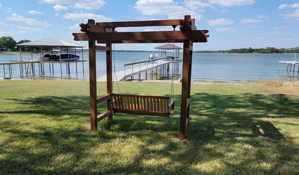 A wooden porch swing on a grassy lawn overlooking the calm water and private boat docks of Eagle Mountain Lake near Azle, TX, perfect for a relaxing outdoor getaway.