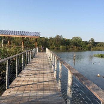 A long wooden boardwalk trail extending over the water at the Fort Worth Nature Center & Refuge, surrounded by lush green trees and wetlands near Azle, TX.