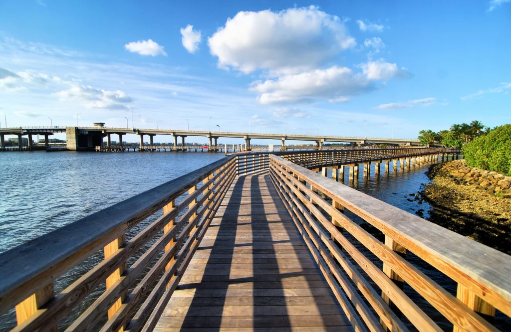 A long, winding wooden boardwalk and fishing pier stretching over the blue water of Lake Worth near Azle, Texas, with a large concrete bridge and white clouds in the background.