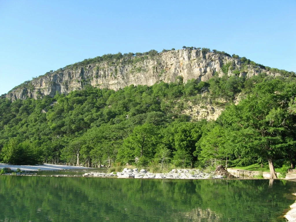 A serene view of the lake at Daingerfield State Park, featuring a swimming platform on the blue water, a green lawn with a volleyball net, and vibrant fall foliage on the surrounding pine trees—a top destination for RV road trips from Azle.