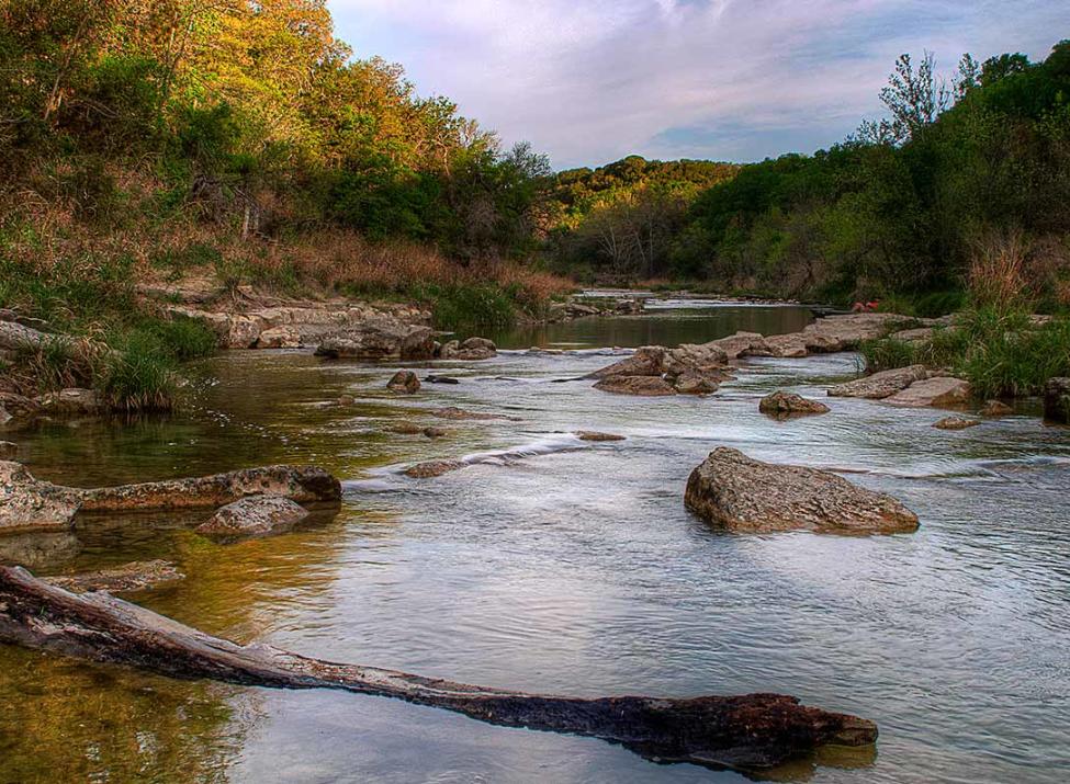 A scenic view of the Paluxy River at Dinosaur Valley State Park, showing clear water flowing over large flat stones and ancient riverbeds surrounded by lush green trees—a popular stop for RV road trips from Azle.