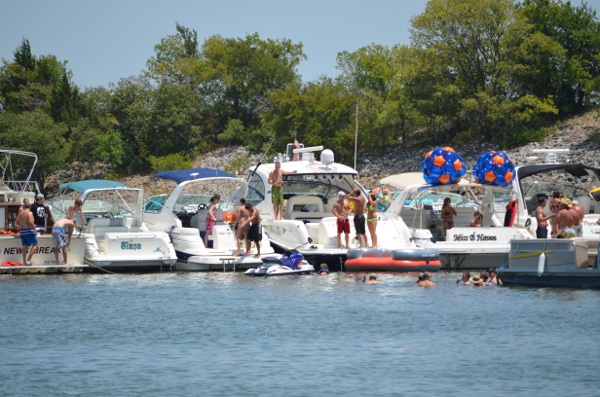A lively scene at Party Cove on Eagle Mountain Lake, featuring multiple boats tied together, people swimming in the water, and colorful orange and blue inflatables on a sunny afternoon—a popular day trip for those RV camping near Fort Worth.