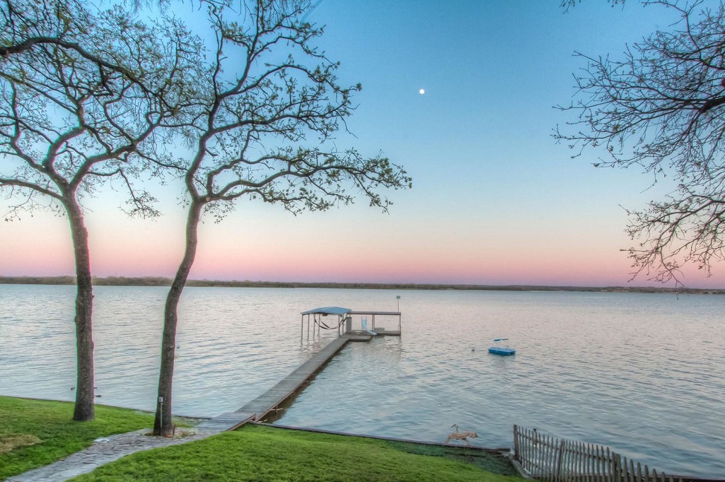A tranquil dusk view of Eagle Mountain Lake, a top destination for RV camping near Fort Worth, featuring a long wooden pier extending into calm water under a soft pink and blue sunset sky with a visible moon.