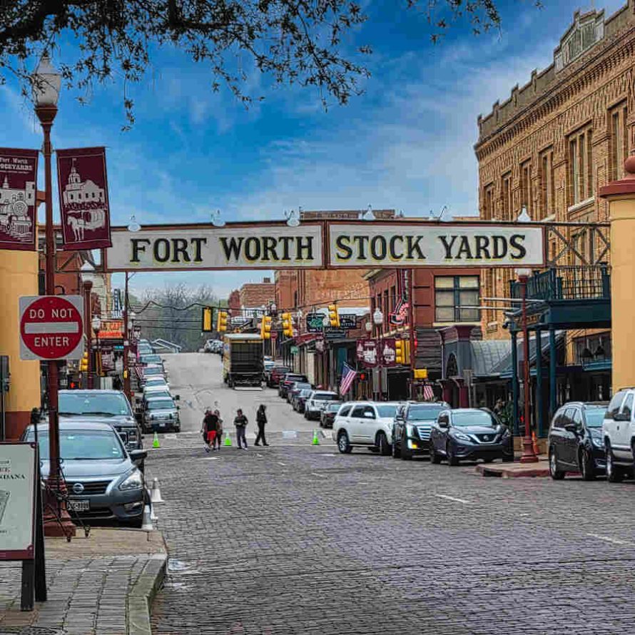 The historic entrance sign of the Fort Worth Stockyards spanning across a cobblestone street, featuring vintage brick architecture, parked vehicles, and a clear blue sky—a must-see destination for travelers RV camping near Fort Worth.