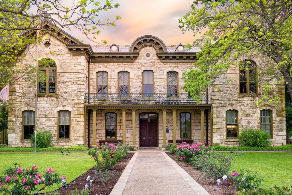 The historic Fredericksburg public library featuring a stunning cream-colored limestone facade, a second-story wrought iron balcony, and dark wooden doors, surrounded by a manicured green lawn and pink rose bushes during a soft sunset.