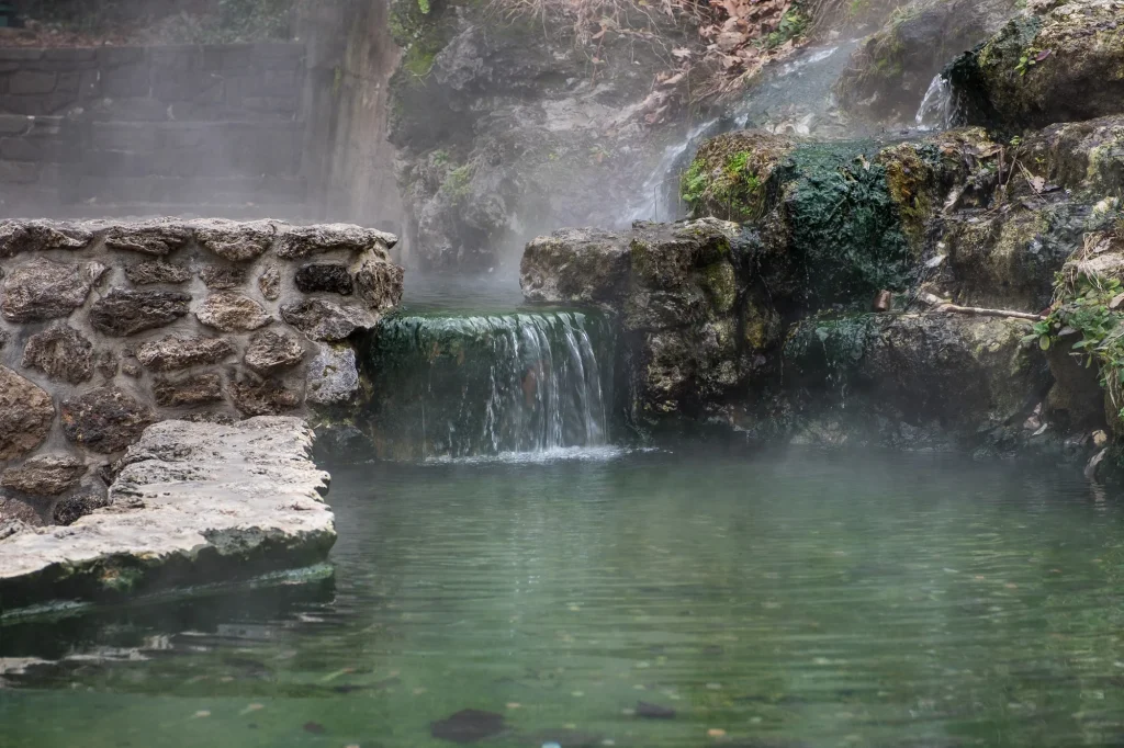 A natural steaming thermal water pool at Hot Springs National Park, featuring a small waterfall flowing over moss-covered stones into a clear green pool, a perfect stop for RV road trips from Azle.