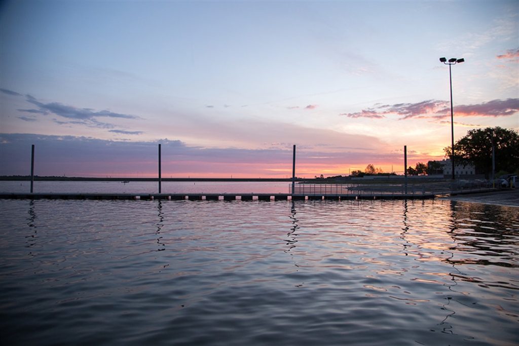 A wide view of Lake Arlington at sunset, featuring a long floating dock on the rippling water, a boat ramp, and a colorful pink and purple sky—a peaceful destination for travelers RV camping near Fort Worth.