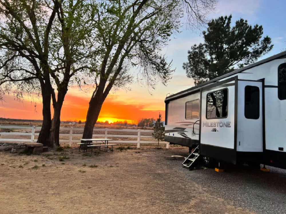 A modern Milestone fifth-wheel trailer parked at a campsite in Azle during a vibrant orange sunset, featuring a white fence, picnic table, and large trees—essential for planning your first RV stay.