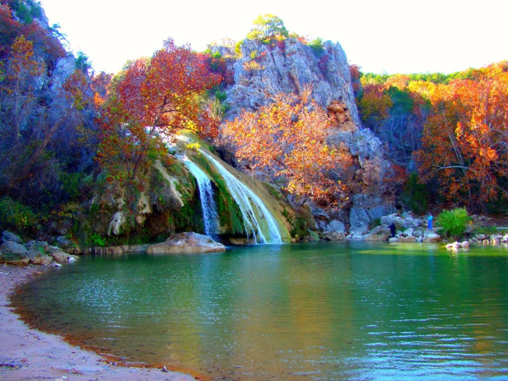 A vibrant landscape view of the main waterfall at Turner Falls Park in Oklahoma, featuring turquoise water, a sandy shoreline, and bright orange and red autumn foliage on the surrounding limestone cliffs—a scenic stop for an RV road trip from Azle.
