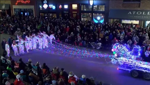 A festive nighttime scene from the Grapevine Parade of Lights, featuring a illuminated parade float pulling performers in glowing white suits covered in LED lights through a crowded street lined with spectators.
